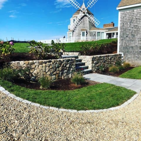 A crushed stone driveway curves around a patch of grass with steps through a retaining wall leading to a windmill